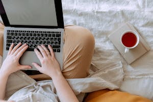 Top view anonymous remote worker typing on keyboard of laptop with blank screen while sitting with crossed legs on bed with tea aside at home