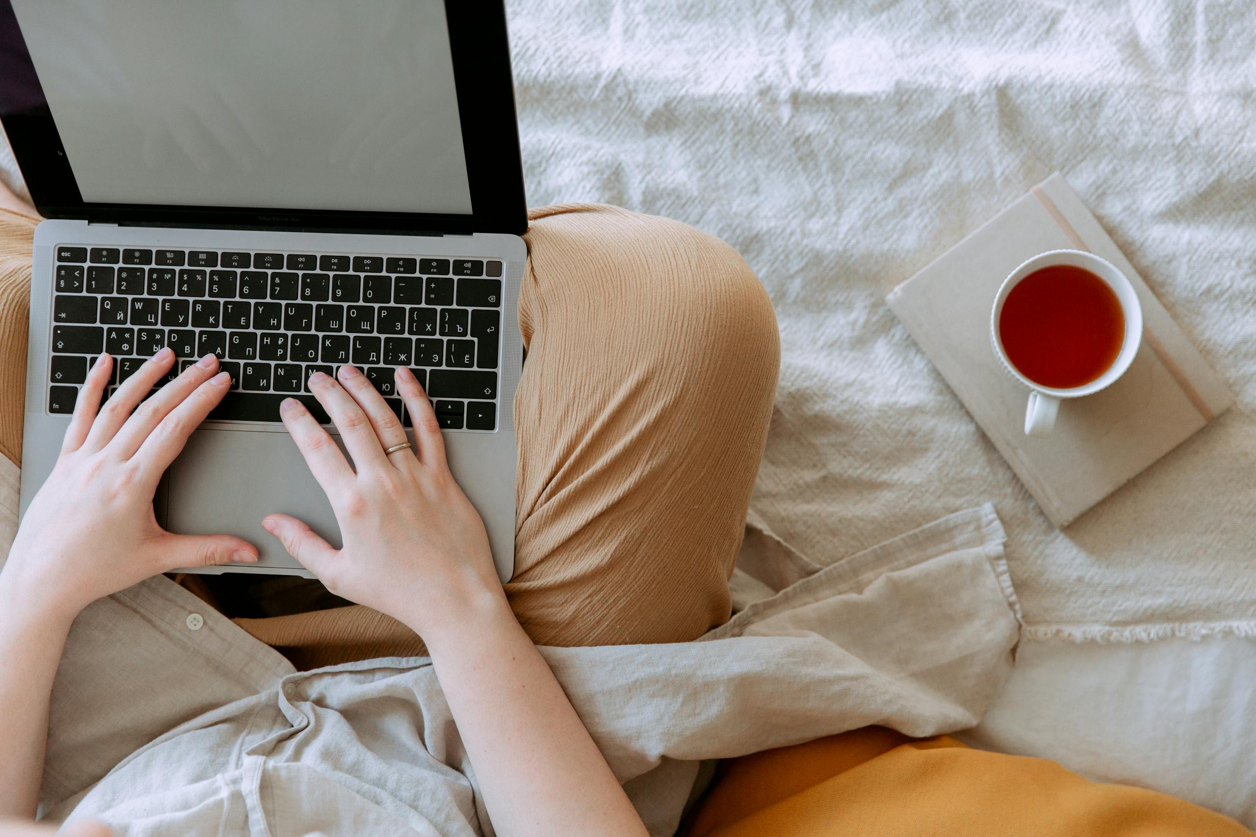 Top view anonymous remote worker typing on keyboard of laptop with blank screen while sitting with crossed legs on bed with tea aside at home