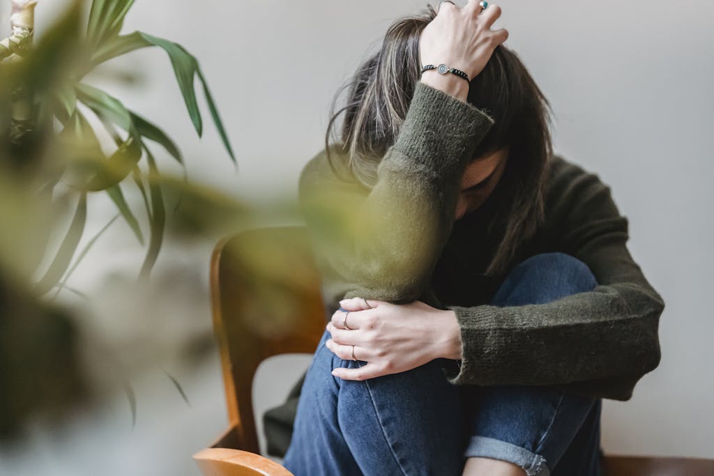 Anonymous young sorrowed female in casual outfit touching dark hair and embracing knees while sitting on chair at home