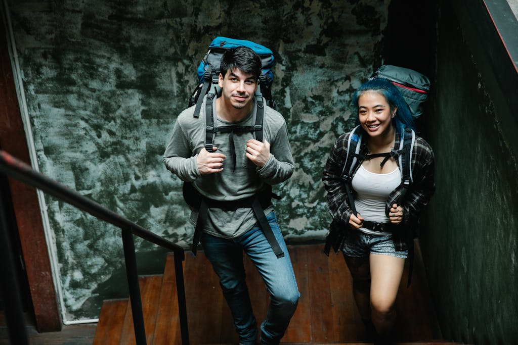 From above of positive diverse couple of travellers with backpacks standing together on narrow wooden staircase