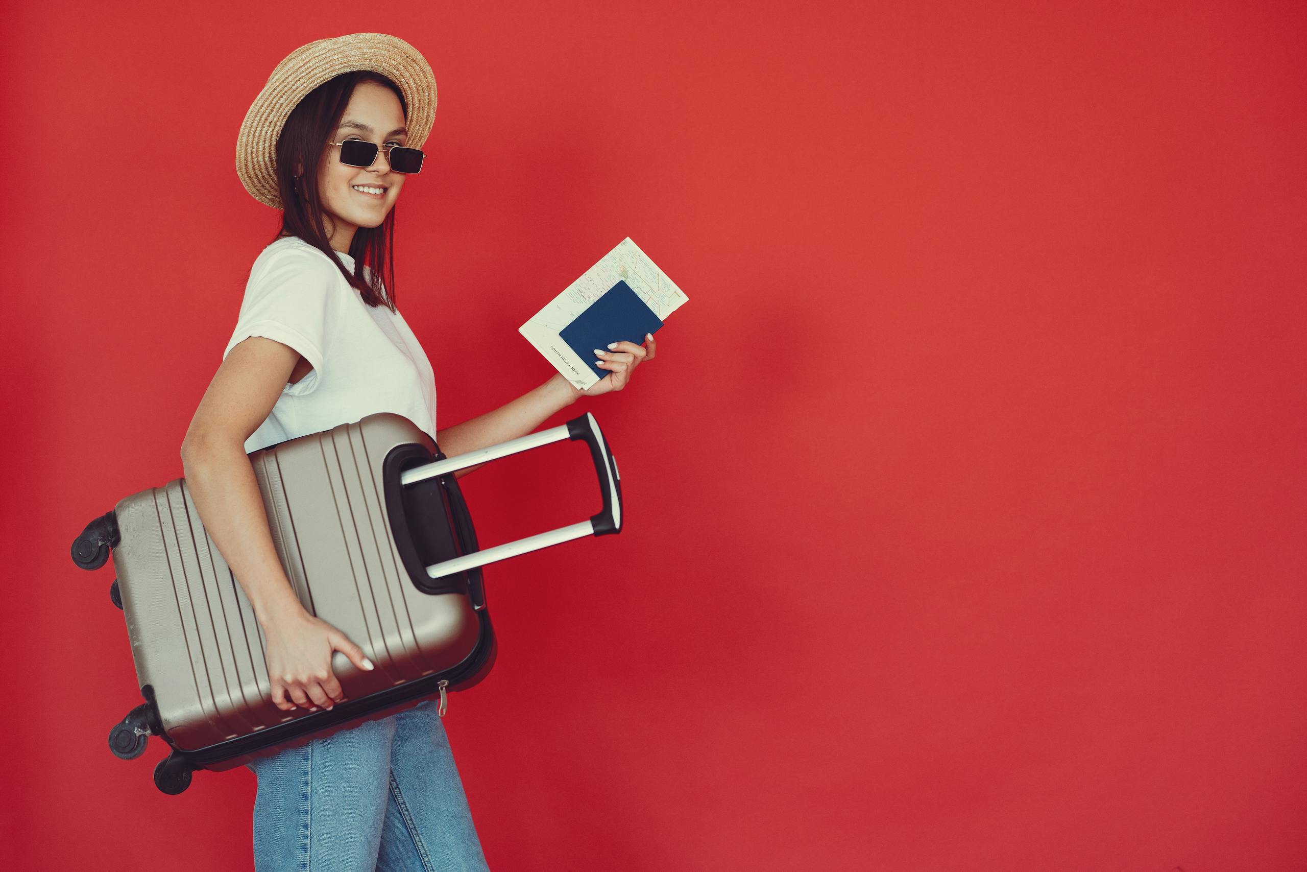 Side view of cheerful young woman with passport and tickets smiling and looking at camera while walking with baggage on red background